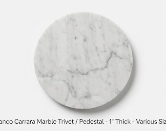May include: A circular Bianco Carrara marble trivet or pedestal with a white base and grey veining. The stone surface is 1 inch thick. The text below the object reads: Bianco Carrara Marble Trivet / Pedestal - 1" Thick - Various Sizes.