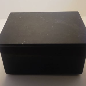 May include: A black rectangular box with a lid. The box is made of a smooth, hard material and has a slightly textured surface.