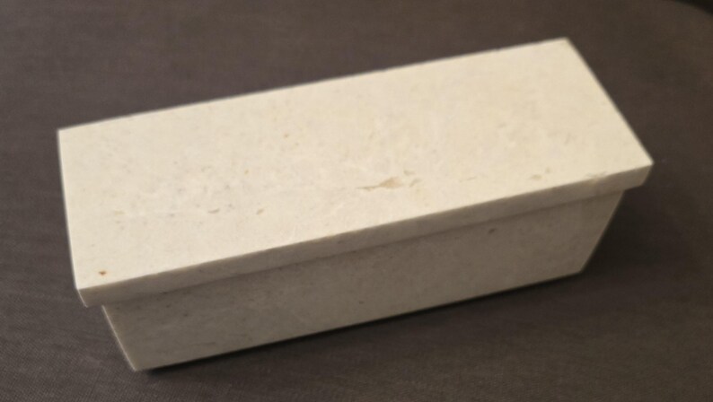 Puede incluir: Una peque&ntilde;a caja rectangular hecha de piedra blanca con una superficie texturizada. La caja tiene una tapa con bisagras y est&aacute; cerrada.