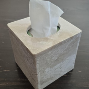 May include: A square tissue box made of beige stone-like material. White tissues are pulled from the circular opening on top. The box has a marbled appearance and sits on a dark wooden surface.