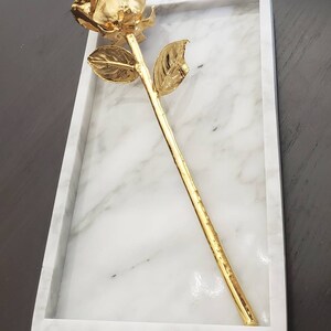 May include: A gold-plated rose on a white marble tray. The rose is a single stem with a single leaf on each side.