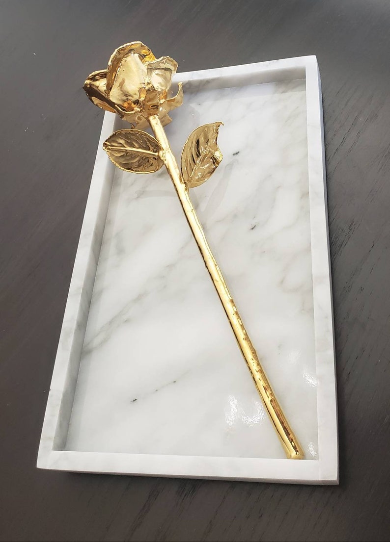 May include: A gold-plated rose on a white marble tray. The rose is a single stem with a single leaf on each side.