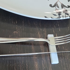 May include: A stainless steel fork and knife rest on a light gray holder, with a matching knife below. A white plate with a black floral design is in the background. The objects are on a dark wooden surface.