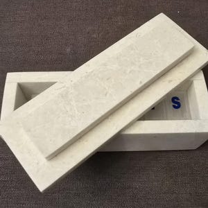 Puede incluir: Una caja de m&aacute;rmol blanco con tapa. La caja tiene forma rectangular y una superficie lisa. La tapa est&aacute; ligeramente entreabierta, revelando el interior de la caja. El interior de la caja tambi&eacute;n es de m&aacute;rmol blanco y tiene una superficie lisa. La caja est&aacute; sobre una superficie marr&oacute;n.