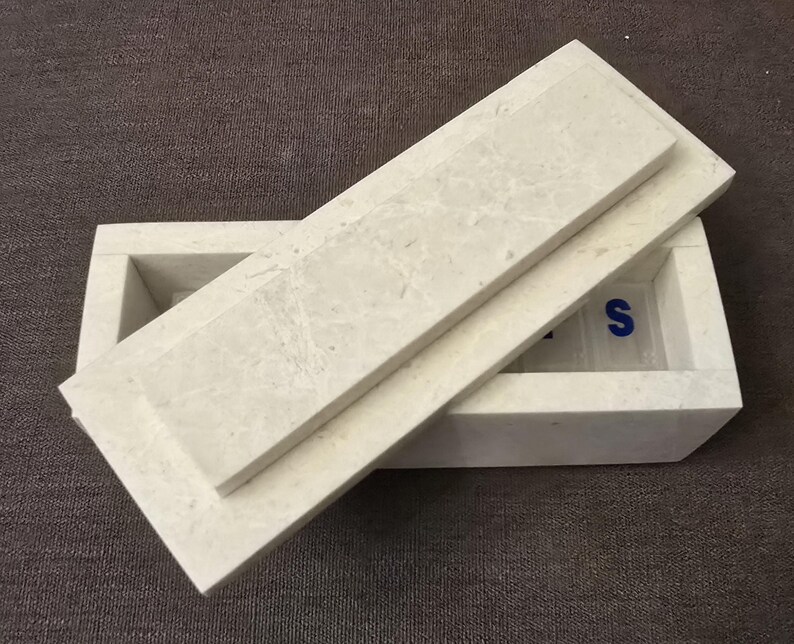 Puede incluir: Una caja de m&aacute;rmol blanco con tapa. La caja tiene forma rectangular y una superficie lisa. La tapa est&aacute; ligeramente entreabierta, revelando el interior de la caja. El interior de la caja tambi&eacute;n es de m&aacute;rmol blanco y tiene una superficie lisa. La caja est&aacute; sobre una superficie marr&oacute;n.