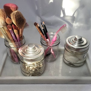 May include: A white marble tray with four glass jars. The jars contain makeup brushes, cotton swabs, cotton balls, and a pink toothbrush.