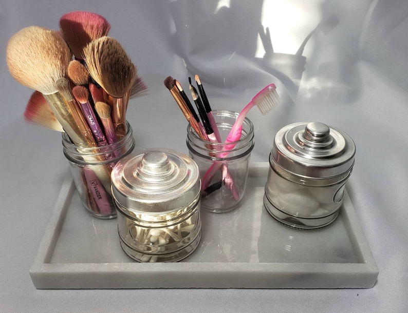 May include: A white marble tray with four glass jars. The jars contain makeup brushes, cotton swabs, cotton balls, and a pink toothbrush.
