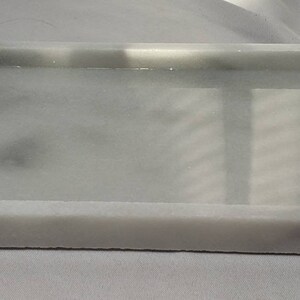 May include: A white rectangular tray with a smooth, slightly textured surface. The tray has a raised rim and is made of a material that resembles marble.
