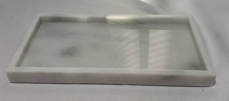 May include: A white rectangular tray with a smooth, slightly textured surface. The tray has a raised rim and is made of a material that resembles marble.