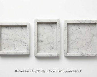 May include: Three rectangular Bianco Carrara marble trays with raised edges and natural grey veining. The trays are displayed in various sizes up to 6 inches by 6 inches by 1 inch. The text below reads: Bianco Carrara Marble Trays – Various Sizes up to 6" x 6" x 1".