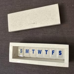 Puede incluir: Pastillero de m&aacute;rmol blanco con tapa. La caja tiene siete compartimentos etiquetados con los d&iacute;as de la semana: S, M, T, W, T, F, S.