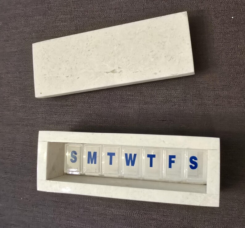 Puede incluir: Pastillero de m&aacute;rmol blanco con tapa. La caja tiene siete compartimentos etiquetados con los d&iacute;as de la semana: S, M, T, W, T, F, S.