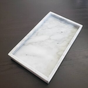 May include: A white marble rectangular tray with a raised rim. The tray is made of natural stone and has a smooth, polished finish.