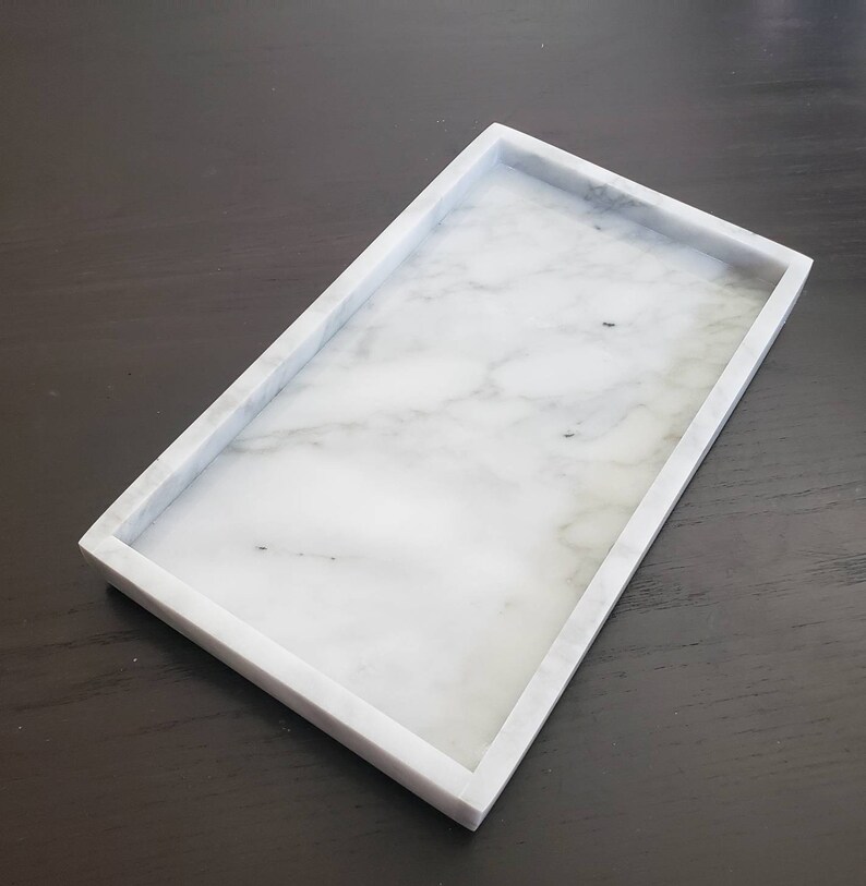 May include: A white marble rectangular tray with a raised rim. The tray is made of natural stone and has a smooth, polished finish.