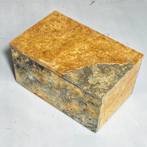 May include: A rectangular box with a hinged lid. The box is made of a stone-like material with a mottled pattern of tan, gray, and black. The lid has a lighter tan color than the sides. The box is likely used for storage or display.
