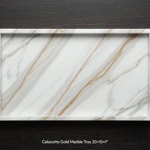 May include: A rectangular Calacatta Gold marble tray, measuring 20 inches long, 10 inches wide, and 1 inch high. The tray features a white background with gold and gray veining, and a raised border.