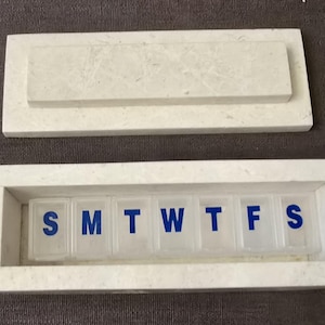 Puede incluir: Pastillero de m&aacute;rmol blanco con tapa. El pastillero tiene siete compartimentos etiquetados con los d&iacute;as de la semana: S, M, T, W, T, F, S.