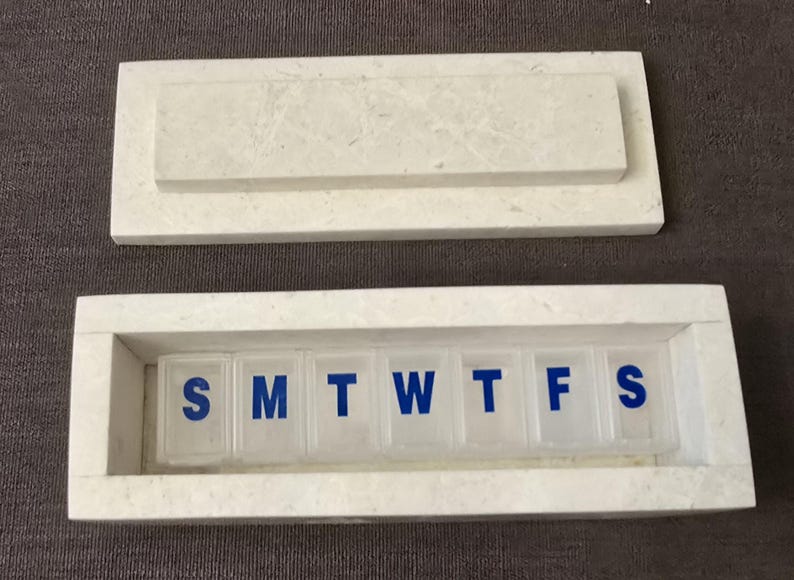 Puede incluir: Pastillero de m&aacute;rmol blanco con tapa. El pastillero tiene siete compartimentos etiquetados con los d&iacute;as de la semana: S, M, T, W, T, F, S.