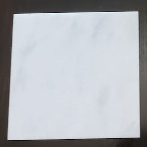 May include: A square, white marble tile with subtle grey veining. The tile has a smooth, polished surface and is set against a dark background. Suitable for various home decor projects.