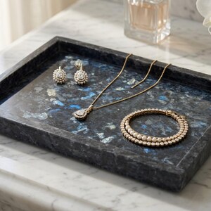 May include: A rectangular tray crafted from dark blue and black stone with a mottled, crystalline texture. The tray holds a gold-toned necklace with a teardrop pendant, a pair of matching stud earrings, and a circular bracelet with white beads.