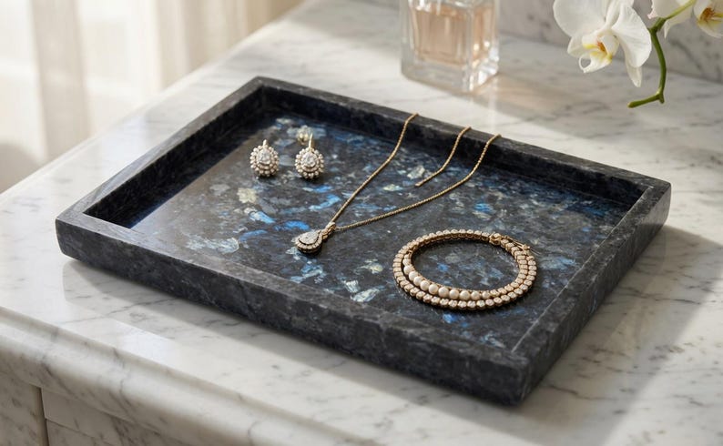 May include: A rectangular tray crafted from dark blue and black stone with a mottled, crystalline texture. The tray holds a gold-toned necklace with a teardrop pendant, a pair of matching stud earrings, and a circular bracelet with white beads.