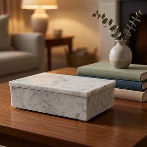 May include: A rectangular white marble box with grey veining and a matching lid. The box is positioned on a wooden surface next to a stack of three hardcover books in shades of green, blue, and beige, topped with a small ceramic vase containing greenery.