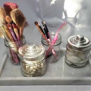 May include: Four glass jars with silver lids on a white marble tray. The jars contain makeup brushes, cotton swabs, cotton balls, and a pink toothbrush.