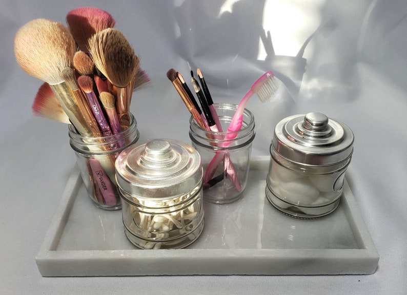 May include: Four glass jars with silver lids on a white marble tray. The jars contain makeup brushes, cotton swabs, cotton balls, and a pink toothbrush.