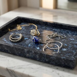May include: A rectangular dark blue stone tray with a mottled texture containing a gold chain necklace with a geometric pendant, a pair of silver hoop earrings, a blue teardrop pendant on a chain, and a silver twisted bangle bracelet.
