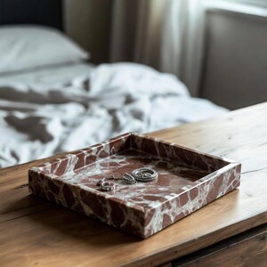 May include: A square tray made of reddish-brown marble with white veining, featuring a raised edge. Inside the tray, there are three small silver-toned rings resting on the smooth, polished stone surface of the decorative container.