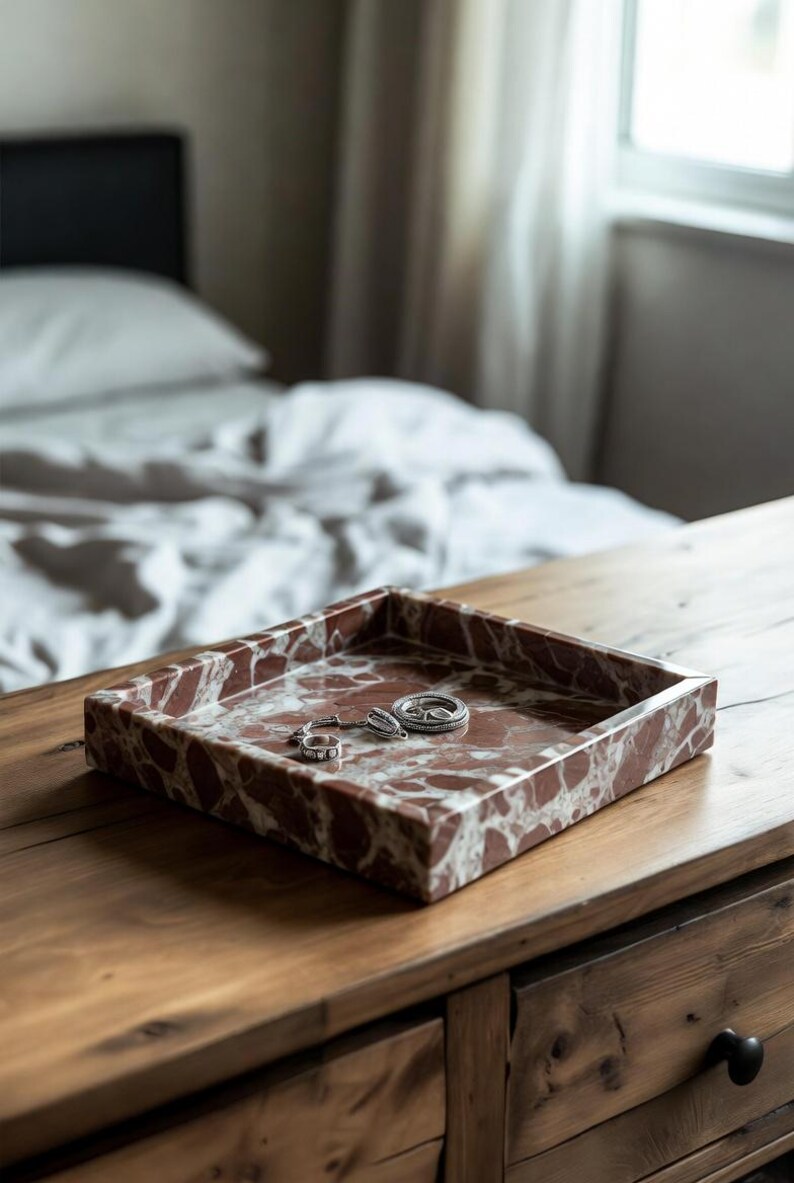 May include: A square tray made of reddish-brown marble with white veining, featuring a raised edge. Inside the tray, there are three small silver-toned rings resting on the smooth, polished stone surface of the decorative container.