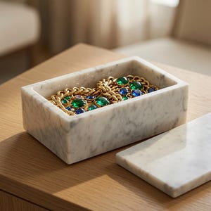 May include: A rectangular white marble box with a matching lid placed to the side. Inside the box rests a gold-toned chain necklace featuring large, faceted emerald green and sapphire blue stones set within the metal links of the jewellery piece.