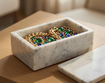 Op de afbeelding: Een rechthoekige doos van wit marmer met een bijpassend deksel dat ernaast ligt. In de doos ligt een goudkleurige ketting met grote, gefacetteerde smaragdgroene en saffierblauwe stenen die in de metalen schakels van het sieraad zijn gezet.