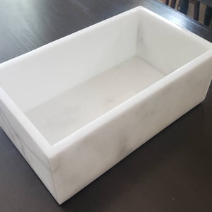 Puede incluir: Una bandeja rectangular de mármol blanco con una superficie lisa y pulida. La bandeja está vacía y lista para ser llenada con artículos decorativos u objetos pequeños.