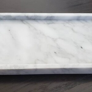 May include: A white marble rectangular tray with a raised rim. The tray has a natural veining pattern.