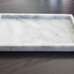 May include: A white marble tray with a rectangular shape and a smooth surface. The tray has a raised edge and is perfect for serving or displaying items.
