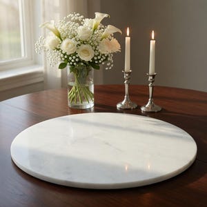 May include: A circular white marble serving board on a dark wood table. A vase of white flowers and two lit candles in silver candlesticks are also on the table. The scene is illuminated by natural light.