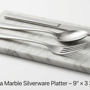 May include: A rectangular Bianco Carrara marble platter measuring 22.86 cm by 9.53 cm, featuring a white surface with grey veining. A stainless steel knife, spoon, and fork are arranged on top of the platter, which is part of a set of 4.