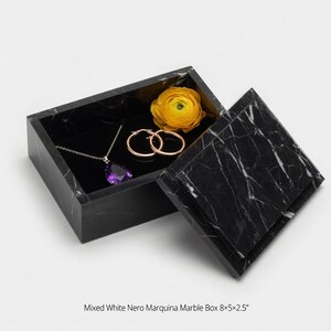 May include: A black marble jewelry box with a white veined pattern, open to reveal a purple pendant necklace, gold hoop earrings, and a yellow flower. The box is rectangular, with the lid slightly ajar.