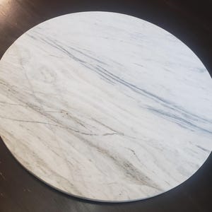 May include: A circular marble tabletop featuring a white and grey veined design. The stone surface is set against a dark brown background, highlighting the natural patterns. The tabletop is a smooth, solid surface.