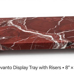 May include: A rectangular Rosso Levanto marble display tray featuring deep red tones with prominent white veining, supported by four short matching marble risers. The text below reads: Rosso Levanto Display Tray with Risers • 8" x 4" x 3/4".