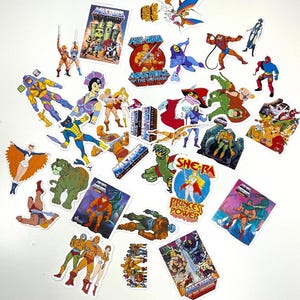 May include: A selection of colourful stickers featuring characters and logos from the "Masters of the Universe" franchise. The stickers showcase various heroes, villains, and scenes, with vibrant colours and detailed artwork. The stickers are scattered on a white surface.