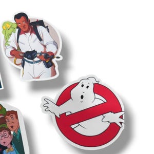 May include: A collection of Ghostbusters-themed stickers. One sticker features a character in a white jumpsuit with a proton pack and a green ghost. Another shows the iconic Ghostbusters logo with a white ghost inside a red circle with a red slash.