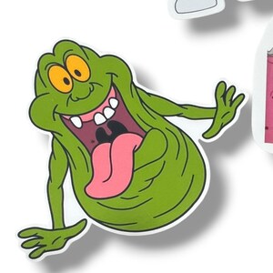 May include: A green cartoon sticker of a smiling, blob-like character with orange eyes, a pink tongue, and white teeth. The character is in a dynamic pose with arms outstretched. The sticker has a white border and is likely from the Ghostbusters franchise.
