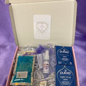 Puede incluir: Una caja de regalo llena de artículos de cuidado personal, que incluyen una bomba de baño azul, una pequeña botella de spray, un amuleto de corazón plateado y dos cajas de té orgánico Pukka night time. La caja está forrada con papel blanco triturado. La tarjeta en la parte superior dice "Todo lo que necesitas es amor".