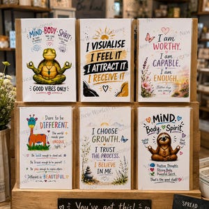 May include: Six rectangular greeting cards displayed on a wooden stand. The cards feature illustrations of a meditating frog, a sloth, a giraffe, and motivational text including phrases like I Visualise, I Choose Growth, and I Am Worthy.