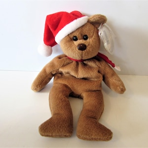 May include: A brown teddy bear wearing a red Santa hat with a white pom-pom and a red scarf. The bear has a brown nose and black eyes. The bear is sitting on a white surface.