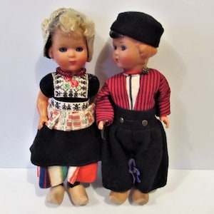 May include: Two vintage Dutch dolls wearing traditional clothing. The girl doll is wearing a blue and white dress with a red and white apron. The boy doll is wearing a red and white striped shirt with black pants. Both dolls have wooden shoes.