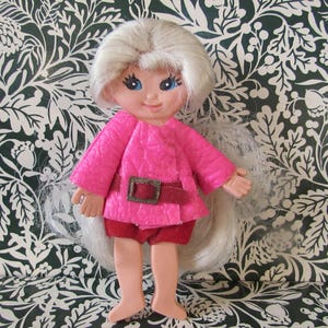 May include: A doll with long white hair wearing a pink jacket with a brown belt and red pants. The doll is standing on a white and black floral patterned background.