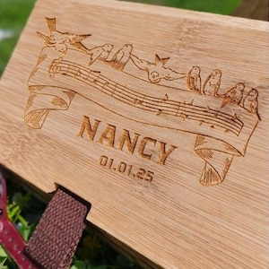 May include: A wooden box with a laser-etched design featuring birds perched on musical notes. The name "NANCY" and the date "01.01.25" are also engraved on the box. The box has a brown strap and is set against a blurred green background.